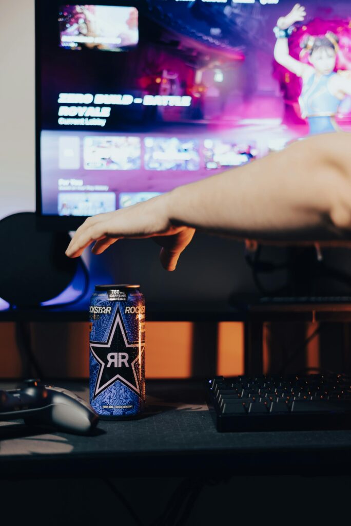 A gamer reaches for an energy drink on a desk with gaming setup and screen.